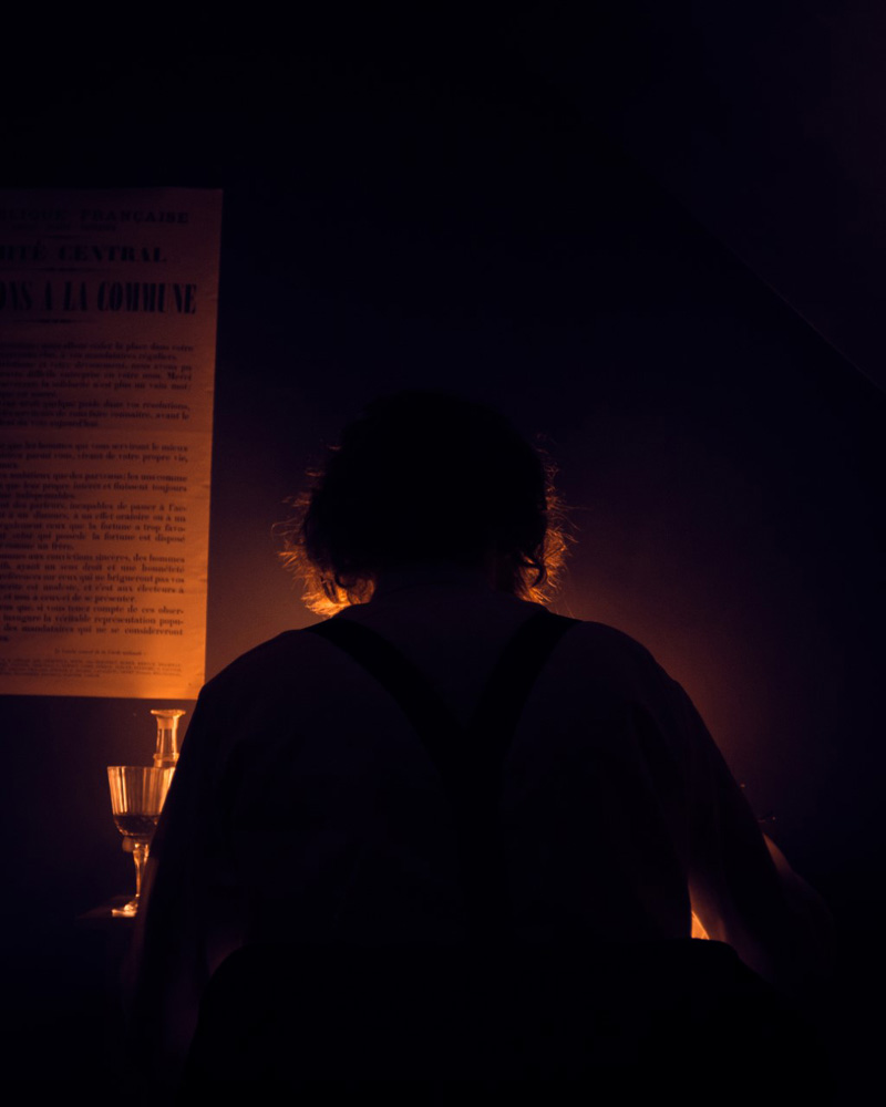 photo dans la pénombre d'une personne assise vue de dos et en buste, seulement éclairée d'une bougie. A gauche, une affiche et un verre